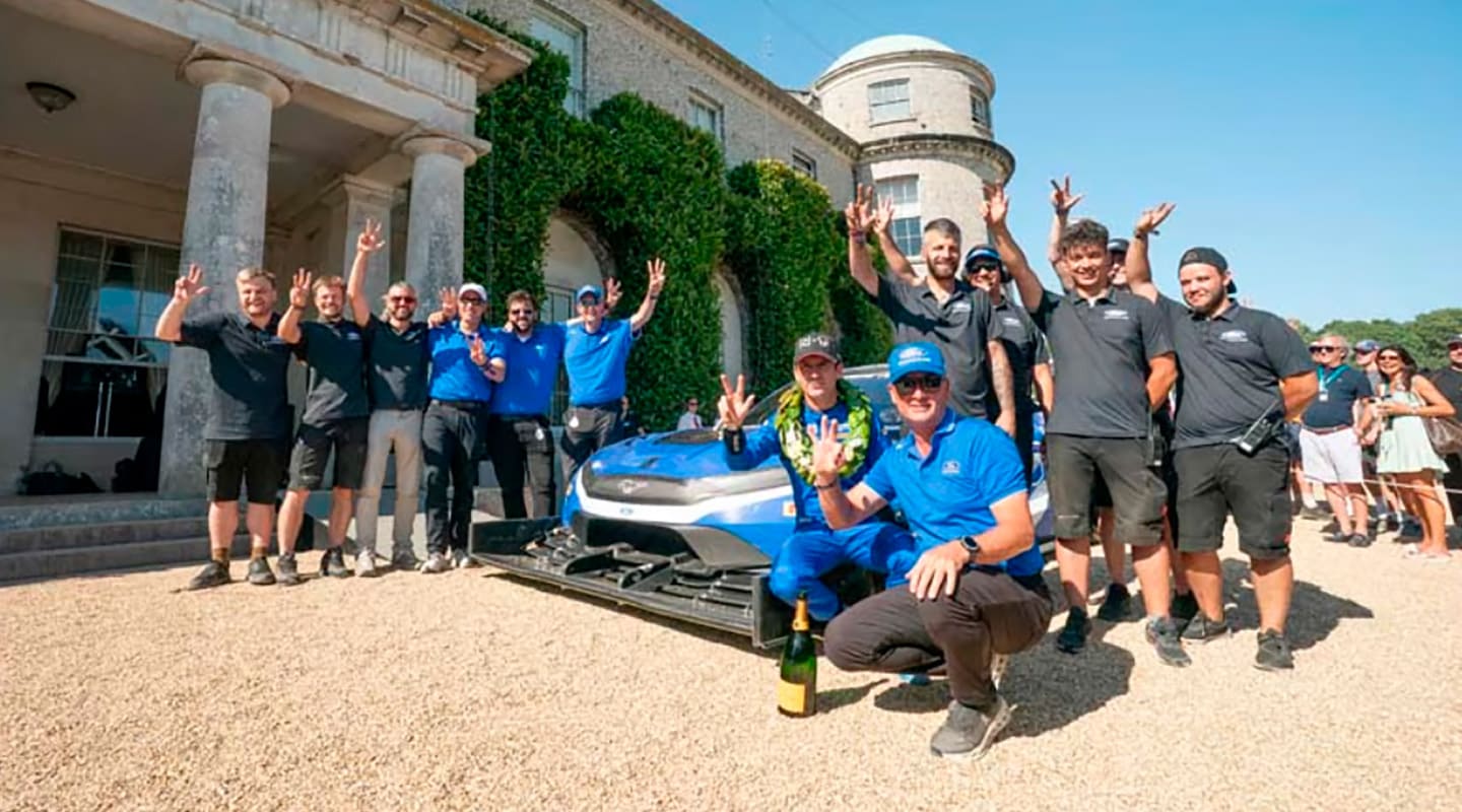 Grupo de personas reunido alrededor de un auto de carreras azul frente a un edificio histórico.