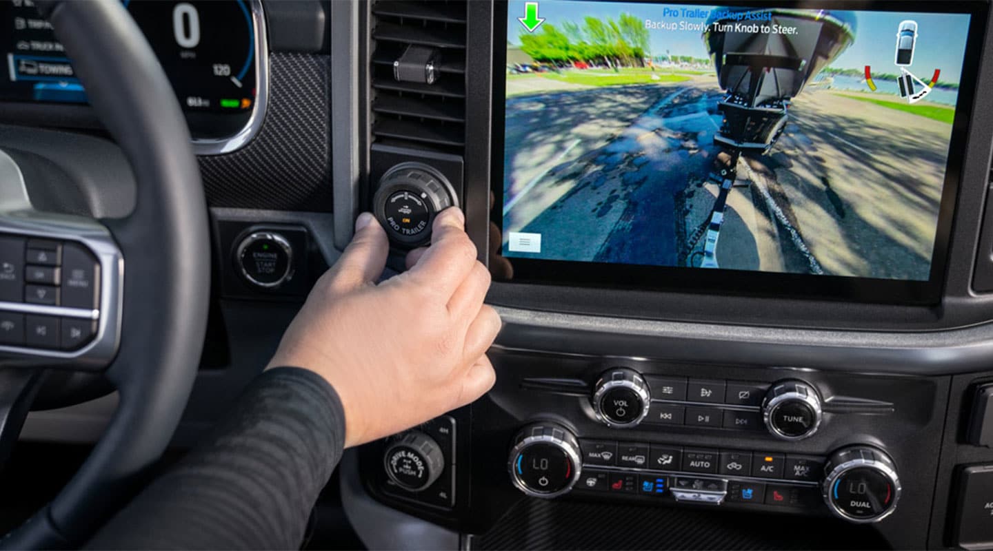 Se muestra en la imagen la amplia pantalla de la cabina de Ford F-150 mientras la mano de un conductor realiza ajustes con una de las perillas